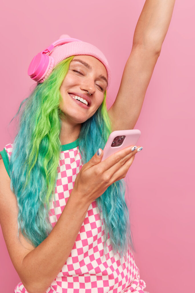 woman with dyed colorful hair dances with arm raised up listens favorite music via headphones holds mobile phone isolated pink